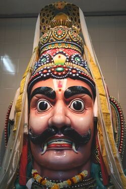 A big moustached male head, with big eyes, big ears and thick eyebrows. Fangs protrude from the sides of his mouth. The head wears a conical crown, with a cobra hood at the top. A floral garland and gold necklace are seen around the neck.